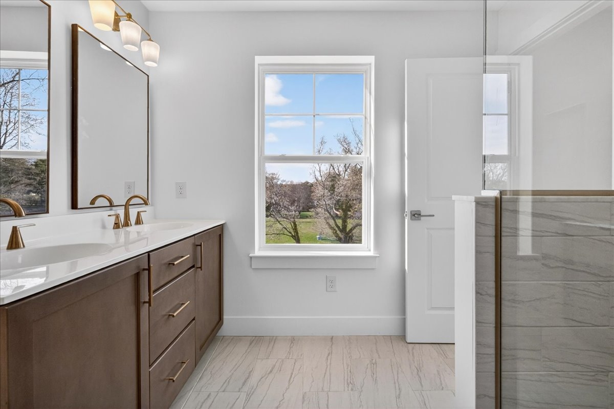 Primary bath double vanity with walnut cabinets, quartz counter, brass fixtures, and a large window overlooking blossoming trees